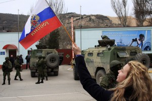 A woman waves a Russian flag as armed servicemen wait near Russian military vehicles outside a Ukrainian border guard post in the Crimean town of Balaclava
